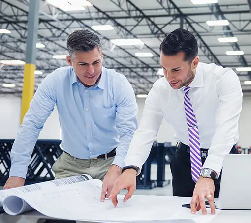 Two men in professional attire reviewing architectural blueprints inside a large industrial warehouse facility.